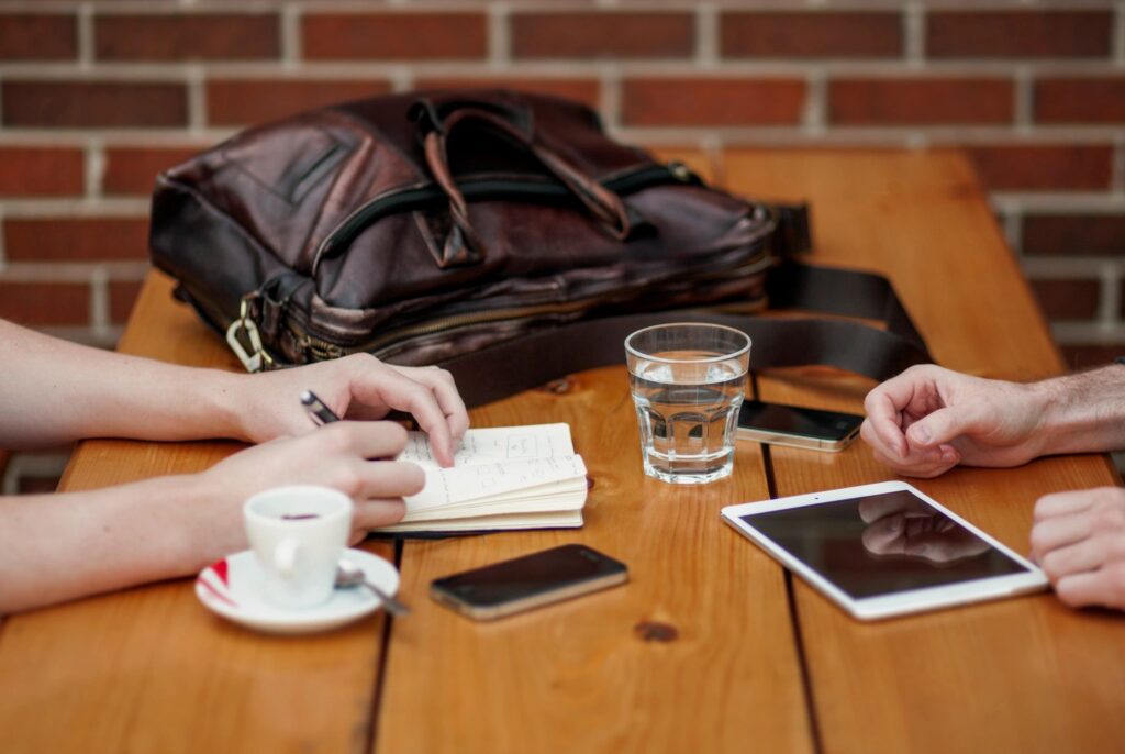 Two people meeting at a table with one note taking, an ipad, water, an iphone on the table 